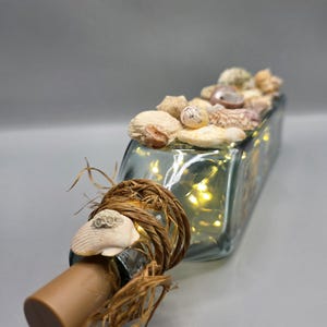 May include: A rectangular glass bottle filled with seashells and small fairy lights. The bottle's neck is adorned with a brown cork, rope, and a seashell. The bottle is filled with small, warm-toned lights.