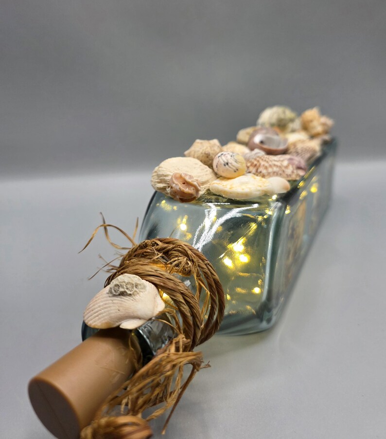 May include: A rectangular glass bottle filled with seashells and small fairy lights. The bottle's neck is adorned with a brown cork, rope, and a seashell. The bottle is filled with small, warm-toned lights.