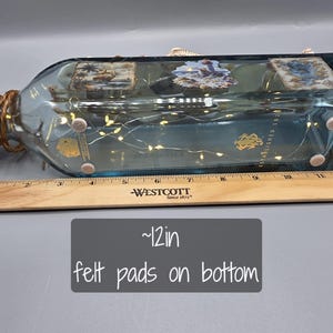 May include: A decorative, rectangular glass bottle with a blue tint, adorned with small lights and nautical-themed images. The bottle is approximately 12 inches long and has felt pads on the bottom.