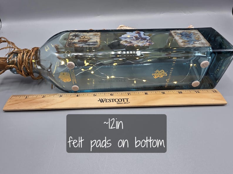 May include: A decorative, rectangular glass bottle with a blue tint, adorned with small lights and nautical-themed images. The bottle is approximately 12 inches long and has felt pads on the bottom.