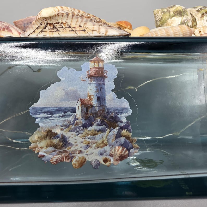 May include: A rectangular, teal-colored glass tray with a painted coastal scene featuring a lighthouse, rocks, and ocean waves. The tray is decorated with various seashells along the top edge.