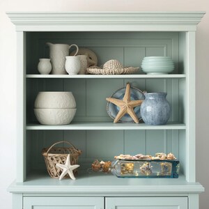 May include: A light blue cabinet with open shelves displays coastal decor. Items include white ceramic pitchers and bowls, a woven hat, a starfish, a blue vase, and a glass bottle filled with shells and lights. The cabinet has a classic design.