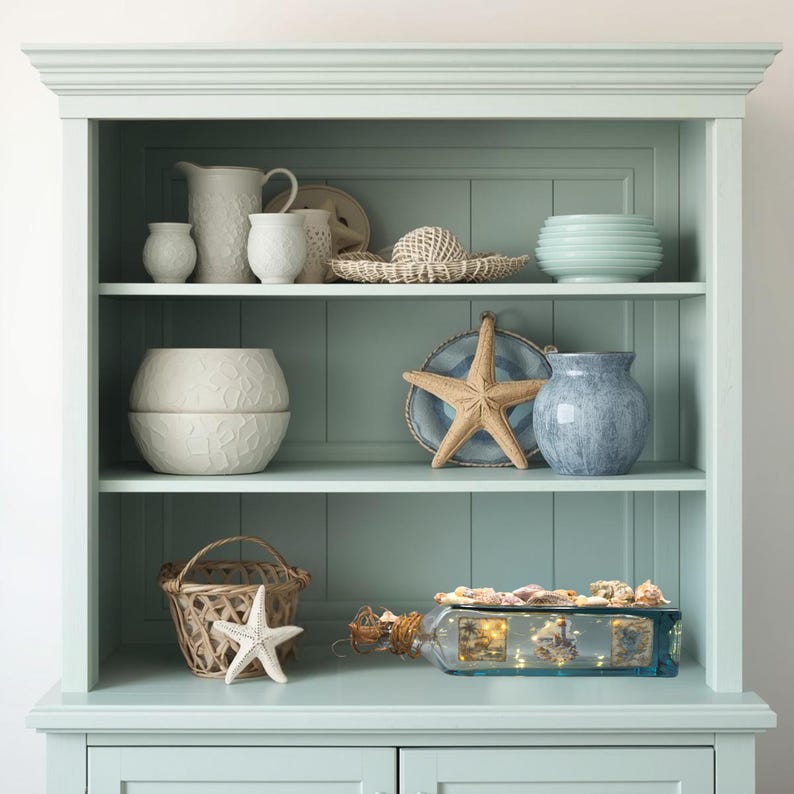 May include: A light blue cabinet with open shelves displays coastal decor. Items include white ceramic pitchers and bowls, a woven hat, a starfish, a blue vase, and a glass bottle filled with shells and lights. The cabinet has a classic design.