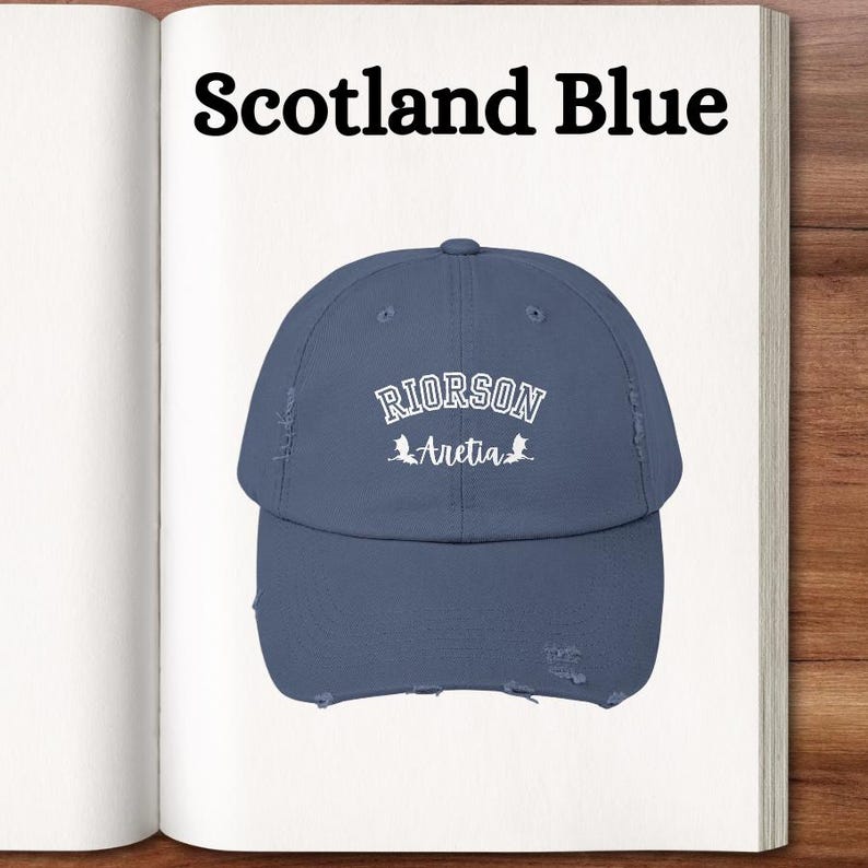 Op de afbeelding: Een distressed, donkerblauwe baseballpet met witte geborduurde tekst die "RIORSON Aretia" luidt en twee kleine witte draken-silhouetten.