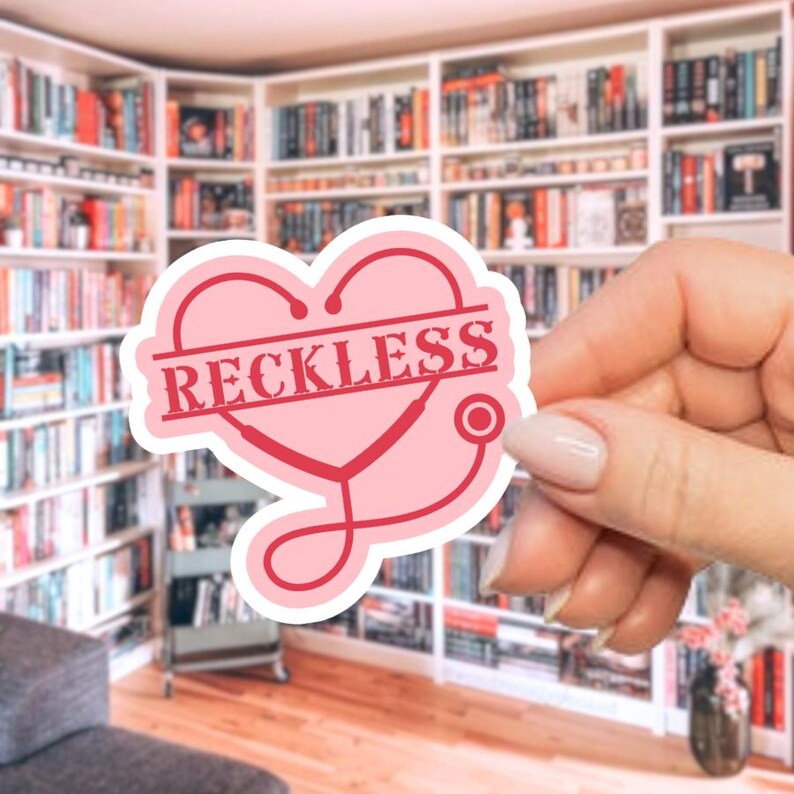 May include: A pink sticker with a heart-shaped design and the word "RECKLESS" in a rectangular stamp. A stethoscope is incorporated into the heart shape. The sticker is held in front of a blurred background of bookshelves.