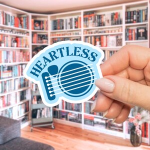 May include: A light blue sticker with the word "HEARTLESS" in a curved font. The sticker features a guitar design with white strings and a light blue outline. The sticker is held in front of a bookshelf.