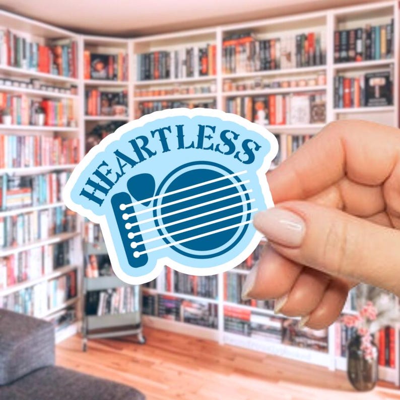 May include: A light blue sticker with the word "HEARTLESS" in a curved font. The sticker features a guitar design with white strings and a light blue outline. The sticker is held in front of a bookshelf.