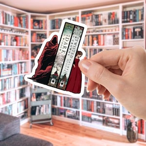 Puede incluir: Una pegatina con tres lomos de libros con los títulos "Dark Window", "One Crowns" y "Twisted Crowns". La pegatina presenta ilustraciones de una figura encapuchada y una mujer con un vestido rojo. El fondo es una biblioteca.