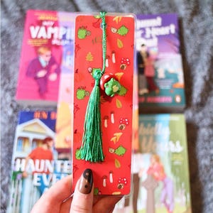Könnte beinhalten: Ein rotes Lesezeichen mit einer grünen Quaste und einem Frosch-Anhänger. Das Lesezeichen hat ein Muster aus grünen Fröschen, Pilzen und Blättern. Mehrere Bücher sind im Hintergrund.