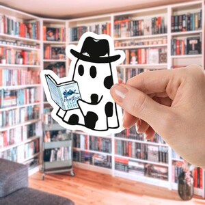 May include: A white sticker featuring a cartoon ghost wearing a black hat and reading a book titled "Heartless" by Elsie Silver. The ghost has black spots and is holding the book in its hands. The background is a library.