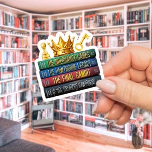 May include: A sticker featuring four books from "The Inheritance Games" series, with titles visible. The sticker includes a gold crown, key, and chess piece. The books are stacked, with green, blue, red, and black spines. The background shows a bookshelf.