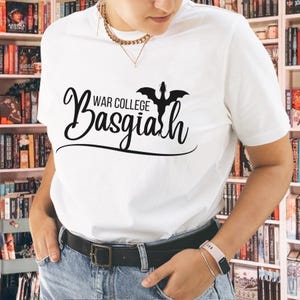 May include: A white short-sleeved t-shirt featuring black text that reads War College Basgiath with a black dragon silhouette above the word Basgiath. The shirt is paired with blue denim jeans and a black belt with a rectangular buckle.
