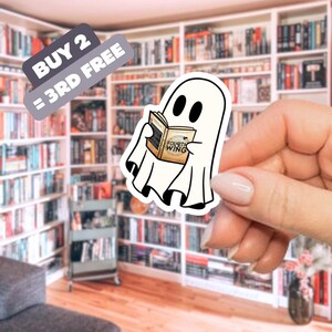 May include: A white ghost sticker reading a book titled "Fourth Wing." The sticker is held in front of a bookshelf. A banner above the sticker reads "BUY 2 = 3RD FREE."