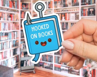 Op de afbeelding: Een blauwe sticker met de tekst "HOOKED ON BOOKS" en een lachend boekpersonage. De sticker heeft een vishaakontwerp en een lint. De achtergrond toont een bibliotheek met veel boeken.