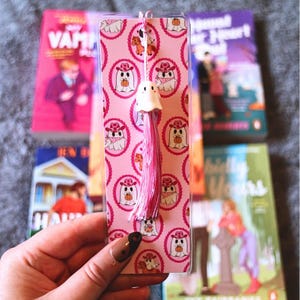 May include: A pink bookmark with a repeating pattern of ghost characters in cowboy hats and holding pumpkins. The bookmark has a pink tassel and a small white ghost charm. Several books are visible in the background.