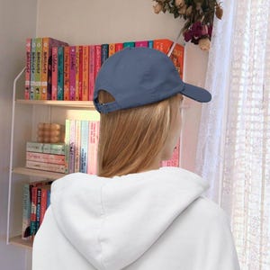 Op de afbeelding: Een persoon draagt ​​een blauwe baseballpet met een witte hoodie. De persoon staat voor een boekenkast vol boeken.