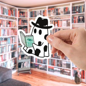 May include: A white sticker featuring a ghost wearing a black hat and reading a book. The ghost has black spots and is holding a book with the title "Hopeless Elsie Silver". The background shows a library.