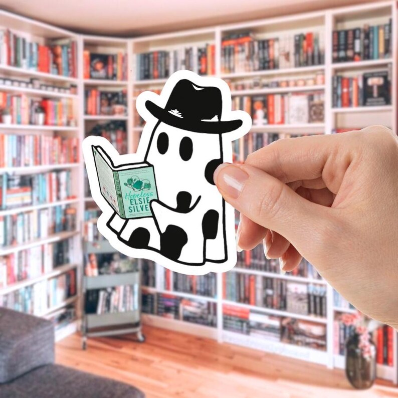 May include: A white sticker featuring a ghost wearing a black hat and reading a book. The ghost has black spots and is holding a book with the title "Hopeless Elsie Silver". The background shows a library.