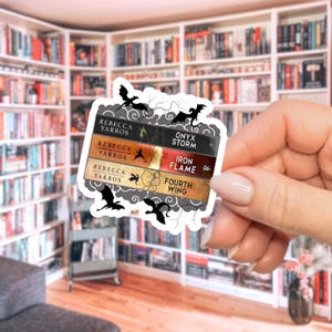 Könnte beinhalten: Ein Aufkleber mit vier Buchrücken mit den Titeln "Onyx Storm", "Iron Flame" und "Fourth Wing" von Rebecca Yarros. Der Aufkleber hat einen weißen Rand mit schwarzen Drachensilhouetten und Wolkenmotiven. Im Hintergrund ist ein Bücherregal zu sehen.