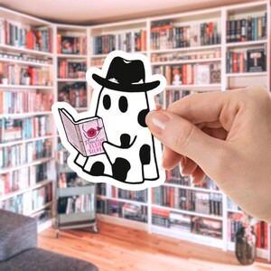 May include: A black and white sticker featuring a cartoon ghost wearing a fedora and reading a book. The book cover reads "Flawless Elsie Silve" with a rose illustration. The ghost has black spots and is set against a blurred bookshelf background.