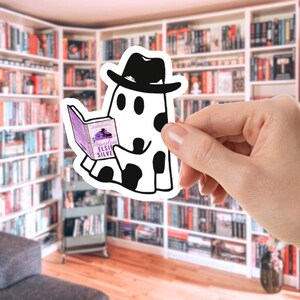 May include: A white sticker featuring a cartoon ghost wearing a black hat and reading a book titled "Powerless" by Elsie Silver. The ghost has black spots and is holding the book with both hands. The background shows a library.