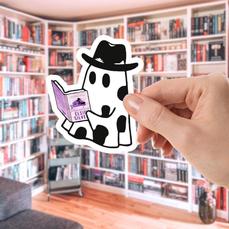 May include: A white sticker featuring a cartoon ghost wearing a black hat and reading a book titled "Powerless" by Elsie Silver. The ghost has black spots and is holding the book with both hands. The background shows a library.