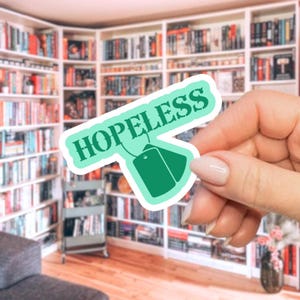 May include: A teal sticker with the word "HOPELESS" in white lettering. The sticker has a tea bag design with two green tags. The background shows a library with many bookshelves filled with books.