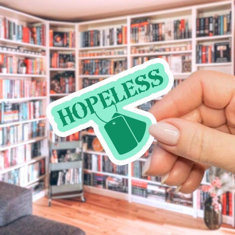 May include: A teal sticker with the word "HOPELESS" in white lettering. The sticker has a tea bag design with two green tags. The background shows a library with many bookshelves filled with books.