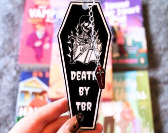 Puede incluir: Marcapáginas negro en forma de ataúd con el texto "DEATH BY TBR". Presenta un esqueleto leyendo un libro, con un dije de ataúd rojo colgante. El fondo borroso muestra libros.