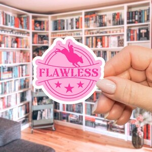 May include: A pink sticker with the word "FLAWLESS" in a circular design. The sticker features a silhouette of a rodeo rider on a bucking bronco, with three stars below the text. The sticker is held in front of a bookshelf.