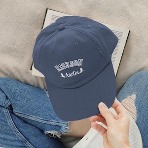 Op de afbeelding: Een marineblauwe baseballpet met witte geborduurde tekst die "RIORSON Aretia" luidt met twee kleine vogelsilhouetten.