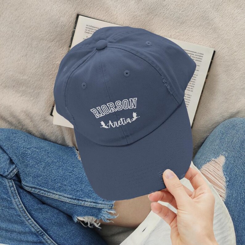 Op de afbeelding: Een marineblauwe baseballpet met witte geborduurde tekst die "RIORSON Aretia" luidt met twee kleine vogelsilhouetten.