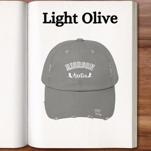 Op de afbeelding: Een licht olijfgroene, verweerde baseballpet met de tekst "RIORSON Aretia" in wit geborduurd.