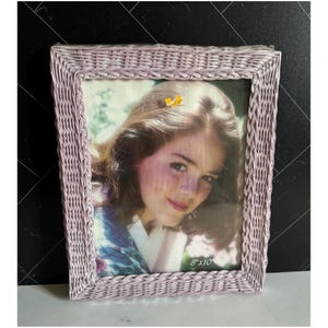 May include: A rectangular, light purple wicker picture frame with a photograph inside. The frame is designed with a woven pattern. The photograph is of a person. The frame is labeled "8"x10".