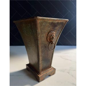 May include: A square-shaped metal planter with a weathered, rusted brown and green patina. The vessel features a flared top, a solid base, and a decorative lion head emblem on the side with a circular metal ring hanging from the lion's mouth.