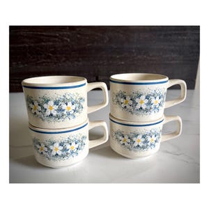 May include: Four ceramic mugs stacked in two pairs. The mugs are off-white with a blue rim and handle. A floral pattern of white and yellow flowers with blue leaves decorates the mugs. The mugs are on a white surface.