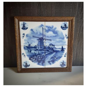 May include: A square wall art piece featuring four ceramic tiles set in a brown wooden frame. The tiles display a blue and white transferware scene of a Dutch landscape with a central windmill, a cow, a small boat, and floral accents.