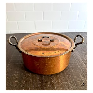 May include: A round copper pot with a matching lid and two curved metal side handles. The exterior of the pot and the lid feature a warm, metallic copper finish with visible signs of natural patina and surface texture throughout the metal.