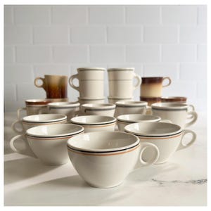 May include: A collection of white ceramic mugs and cups. Some mugs have a gradient brown color, while others are white with black and orange stripes. The cups have a white base with a black and orange stripe design.