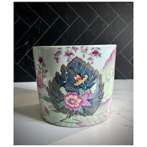 May include: A cylindrical ceramic planter with a floral design. The planter features a light green background with a large blue leaf and pink and purple flowers. Gold accents outline the leaf and flowers.