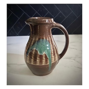 May include: A handmade ceramic pitcher with a brown base and body, featuring a textured design and a teal glaze that drips down the side. The pitcher has a handle and a spout for pouring.