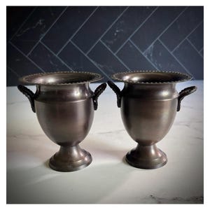 May include: Two bronze-colored metal vases with a classic urn shape. Each vase has a wide, decorative rim, two small handles, and a pedestal base. The vases are displayed on a white surface with a dark blue tiled background.