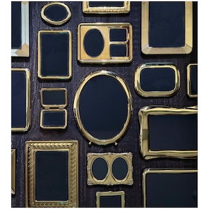 May include: An assortment of gold-colored picture frames in various shapes and sizes, including oval, rectangular, and square. The frames have dark blue or black interiors and are arranged on a dark wood-grain surface.