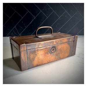 May include: A rectangular, antique copper-toned metal box with a handle and a keyhole. The box has a slightly weathered appearance, with visible patina and signs of age. The handle is a simple, curved design. The box is likely used for storage.