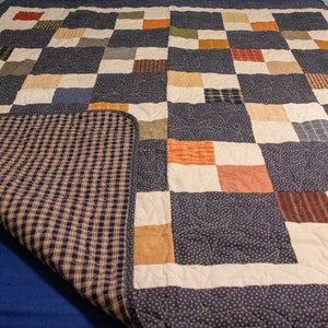 Puede incluir: Una colcha de retazos con un patrón de cuadros azules y blancos. La colcha está hecha de varias telas de diferentes colores y patrones, incluyendo naranja, marrón y beige. La colcha está doblada por la mitad y el lado trasero es visible.