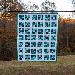 May include: A teal and white quilt with a checkered pattern. The quilt is made up of 36 squares, each with a different design. The quilt is hanging on a black stand in front of a wooded background.