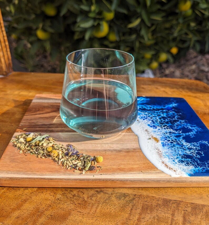 May include: A wooden serving board with a blue and white epoxy resin design resembling ocean waves. A glass of blue herbal tea sits on the board, and loose leaf tea is scattered on the wood.