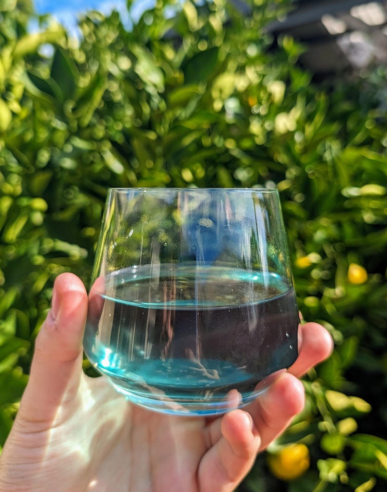 May include: A glass filled with a bright blue liquid, held in a hand against a blurred background of green foliage.