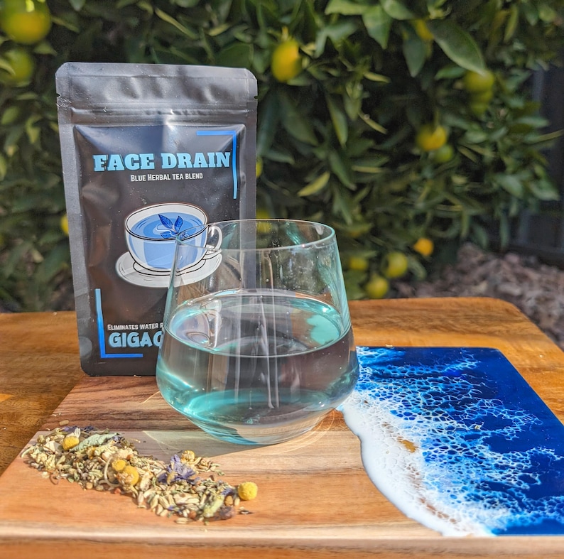 May include: A blue herbal tea blend called "Face Drain" in a black pouch with a glass of blue tea on a wooden board with a blue and white resin design.