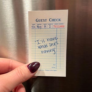 May include: A white guest check with blue lines and the text "GUEST CHECK" printed at the top. The check has a handwritten message that says "I'll have what she's having".